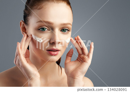Skincare. Ginger-Haired Young Lady Applying Facial Cream Moisturizing And Caring For Skin Standing Posing Over Gray Studio Background. Skin Care Cosmetics For Youth Concept Skincare. Ginger-Haired Young Lady Applying Facial Cream Moisturizing And Caring For Skin Standing Posing Over Gray Studio Background. Skin Care Cosmetics For Youth Concept 134028091