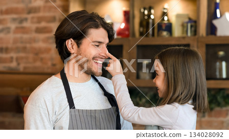 Happy little girl having fun with her dad while cooking together in kitchen, playfully rubs his nose with flour and laughing Happy little girl having fun with her dad while cooking together in kitchen, playfully rubs his nose with flour and laughing 134028092