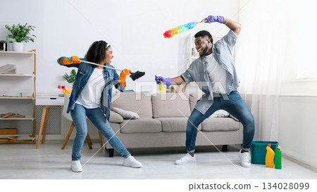 Cleaning Fun. Cheerful Black Couple Fooling While Tidying Up Their Flat, Playfully Fighting With Duster And Swab, Staying At Home, Free Space 134028099