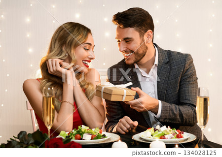 Happy man giving valentine's day present to his surprised girlfriend at romantic dinner in restaurant, pleased woman receiving gift box from boyfriend 134028125