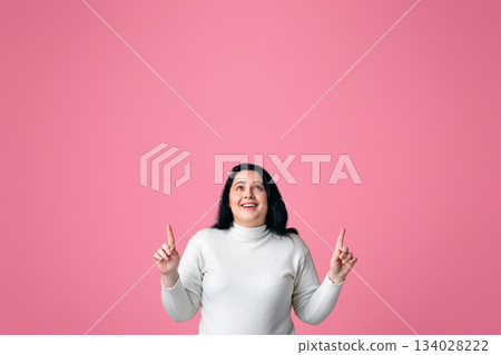 Look Up. Positive Overweight Girl Pointing Fingers Upward Standing Over Pink Studio Background With Copy Space Look Up. Positive Overweight Girl Pointing Fingers Upward Standing Over Pink Studio Background With Copy Space 134028222