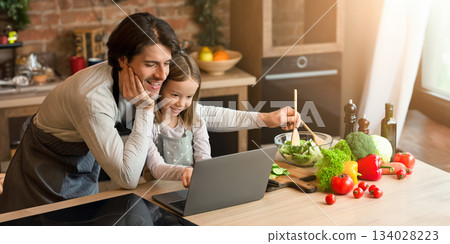 Cheerful single dad and his little daughter using laptop checking ingredients for dinner, cooking in kitchen together Cheerful single dad and his little daughter using laptop checking ingredients for dinner, cooking in kitchen together 134028223