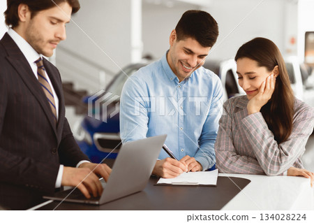 Couple Signing Contract, Warranty Or Guarantee With Car Dealer In Auto Show, Man Filling Application Form Couple Signing Contract, Warranty Or Guarantee With Car Dealer In Auto Show, Man Filling Application Form 134028224