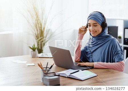 Online Conference Concept. Millennial Arabic Businesswoman Having Video Call With Her Foreign Partners, Using Laptop And Headset In Office Online Conference Concept. Millennial Arabic Businesswoman Having Video Call With Her Foreign Partners, Using Laptop And Headset In Office 134028277