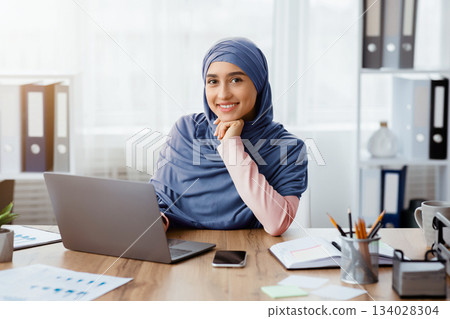 Successful Muslim Businesswoman. Portrait Of Smiling Islamic Female Entrepreneur At Her Workplace In Modern Office, Empty Space Successful Muslim Businesswoman. Portrait Of Smiling Islamic Female Entrepreneur At Her Workplace In Modern Office, Empty Space 134028304