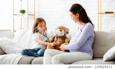 Little girl playing doctor with her pregnant young mom, daughter using stethoscope, treating teddy bear, sitting on sofa at home Little girl playing doctor with her pregnant young mom, daughter using stethoscope, treating teddy bear, sitting on sofa at home 134028311
