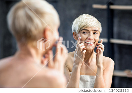 Happy Middle-Aged Woman Applying Facial Cream Moisturizer On Face Standing Wrapped In Towel In Bathroom. Mature Skin Treatment. Selective Focus 134028313