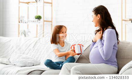Smiling preschool girl giving gift to her pregnant mom, Mother's Day concept, home interior, copy space 134028333