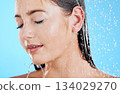 Shower, water and woman face in a studio relax from cleaning and facial skincare. Wellness, splash and beauty routine of female model in bathroom from dermatology and self care with blue background 134029270