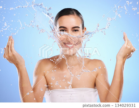 Water splash, portrait or happy woman and face wash for skincare or hygiene on blue background in studio. Smile, wet or model and clean hands in wellness washing, beauty grooming or facial treatment 134029331