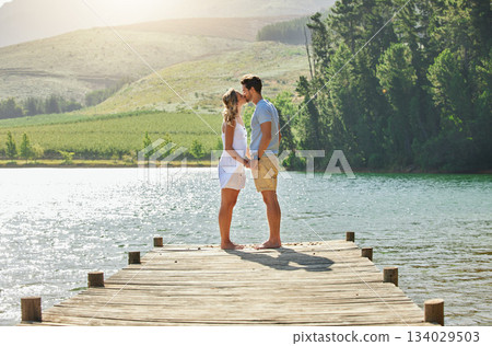 Couple, kissing and holding hands by the lake in romance for love, care and affection together in nature. Man and woman kiss by water on a romantic getaway, summer vacation or date by the lakeside 134029503