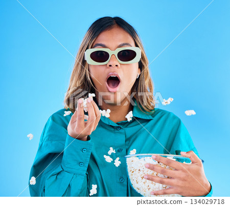 3D movie, popcorn and wow with a woman in studio on a blue background looking shocked while eating a snack. Portrait, glasses and video entertainment with an attractive young female feeling surprised 3D movie, popcorn and wow with a woman in studio on a blue background looking shocked while eating a snack. Portrait, glasses and video entertainment with an attractive young female feeling surprised 134029718