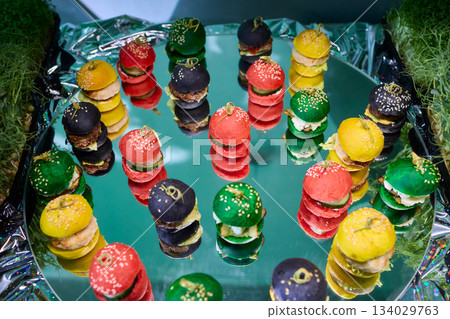 Colorful miniature burger macarons displayed on shiny green foil surface, Playfully arranged rainbow hued miniature burger macarons with glossy shells and festive appeal 134029763