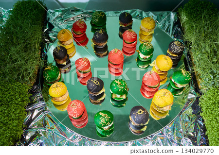 Colorful miniature burger macarons displayed on shiny green foil surface, Playfully arranged rainbow hued miniature burger macarons with glossy shells and festive appeal Colorful miniature burger macarons displayed on shiny green foil surface, Playfully arranged rainbow hued miniature burger macarons with glossy shells and festive appeal 134029770