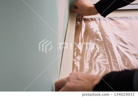 Hand applying masking tape to wall preparing crisp paint edge during interior renovation, closeup of wrist and jacket sleeve, soft natural light, careful steady technique Hand applying masking tape to wall preparing crisp paint edge during interior renovation, closeup of wrist and jacket sleeve, soft natural light, careful steady technique 134029931