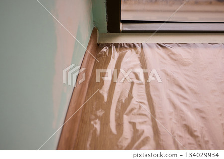 Hand applying masking tape to wall preparing crisp paint edge during interior renovation, closeup of wrist and jacket sleeve, soft natural light, careful steady technique 134029934