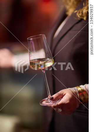 Refined woman savoring amber glass drink, Closeup of woman enjoying sophisticated wine experience, Fashionable lady gracefully holding glass of golden wine at classy Refined woman savoring amber glass drink, Closeup of woman enjoying sophisticated wine experience, Fashionable lady gracefully holding glass of golden wine at classy 134029937