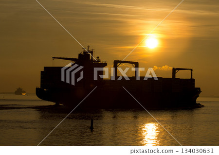 Big containership with cargo sailing towards the sunset in the dusk on the Western Scheldt. Big containership with cargo sailing towards the sunset in the dusk on the Western Scheldt. 134030631