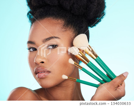 Portrait, brushes and black woman with makeup, foundation and glow against a blue studio background. Face, Nigerian female and lady with cosmetic tools, grooming and morning routine for smooth skin 134030633