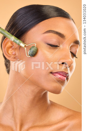 Face, skincare roller and woman with eyes closed in studio isolated on a brown background. Dermatology, facial massage and Indian female model with jade crystal for healthy skin treatment and beauty. 134031020