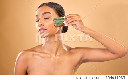 Face, skincare stone and woman with eyes closed in studio isolated on a brown background. Dermatology, facial massage and Indian female model with jade crystal or gua sha for healthy skin treatment. Face, skincare stone and woman with eyes closed in studio isolated on a brown background. Dermatology, facial massage and Indian female model with jade crystal or gua sha for healthy skin treatment. 134031085