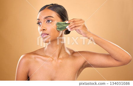 Face, beauty skincare and woman with gua sha in studio isolated on a brown background. Dermatology, thinking and serious Indian female model with jade crystal or stone for healthy skin treatment. 134031086