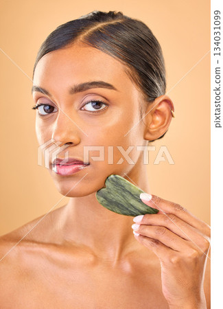 Face portrait, skincare and woman with gua sha in studio isolated on a brown background. Dermatology, massage and serious Indian female model with jade crystal or stone for healthy skin treatment. 134031099
