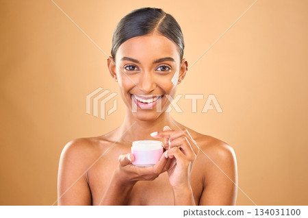 Face, skincare and woman with cream jar in studio isolated on a brown background. Dermatology cosmetics, portrait and happy Indian female apply lotion, creme and moisturizer product for healthy skin. 134031100