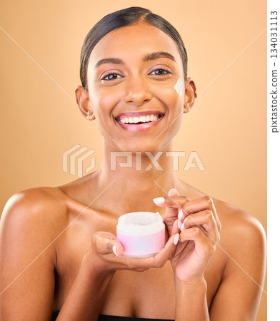 Face, skincare and woman with cream jar in studio isolated on a brown background. Dermatology cosmetics, portrait and happy Indian female apply lotion, creme and moisturizer product for healthy skin. 134031113