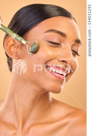 Woman, face and beauty, roller and skincare, smile and facial massage on studio background. Jade quartz cosmetic tools, dermatology and skin glow, happy Indian female and self care with cosmetics 134031243