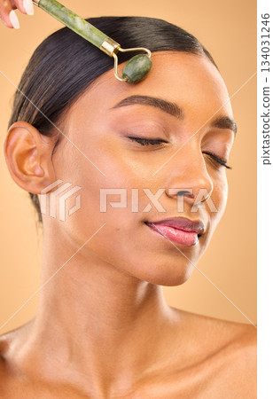 Face, skincare roller and woman with eyes closed in studio isolated on a brown background. Dermatology, facial massage and Indian female model with jade crystal for healthy skin treatment and beauty. 134031246