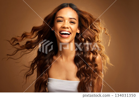 Portrait, hair and wind with an excited woman on a brown background in studio for natural haircare. Face, smile and shampoo with an attractive young female feeling happy about keratin treatment 134031350