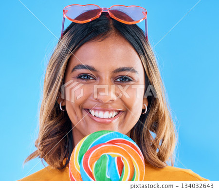 Portrait, lollipop and a woman smiling on a blue background in studio wearing heart glasses for fashion. Face, candy and sweet with a happy young female eating a giant snack while feeling positive Portrait, lollipop and a woman smiling on a blue background in studio wearing heart glasses for fashion. Face, candy and sweet with a happy young female eating a giant snack while feeling positive 134032643
