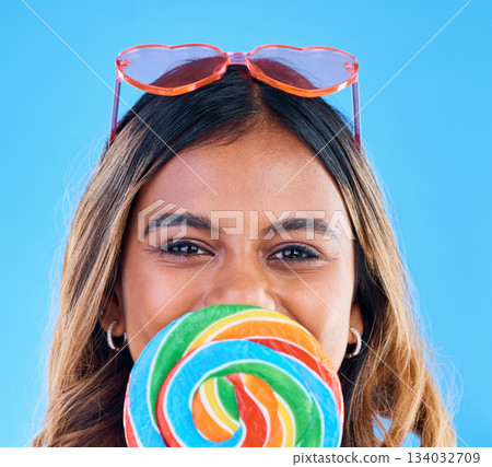 Portrait, giant lollipop and a woman on a blue background in studio wearing heart glasses for fashion. Face, candy and sweet with an attractive young female eating a snack while feeling happy 134032709