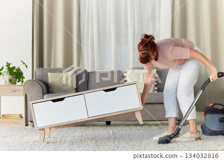 Woman, vacuum and housekeeping on carpet in living room for spring cleaning service, dust or dirt removal at home. Female cleaner lifting table and vacuuming for clean rug, mat or floor in the lounge Woman, vacuum and housekeeping on carpet in living room for spring cleaning service, dust or dirt removal at home. Female cleaner lifting table and vacuuming for clean rug, mat or floor in the lounge 134032816