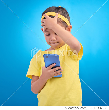 Phone, face palm and a girl on a blue background in studio reading a text message or post on social media. Contact, mobile and mistake with a young female child looking at bad news on her smartphone Phone, face palm and a girl on a blue background in studio reading a text message or post on social media. Contact, mobile and mistake with a young female child looking at bad news on her smartphone 134033262