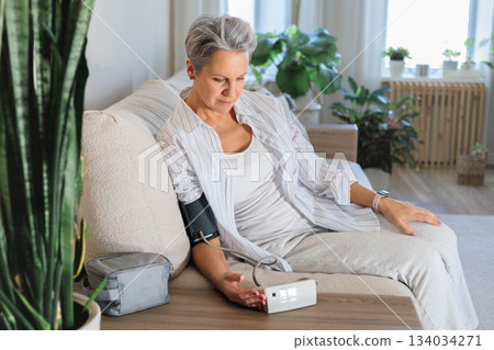 An elderly woman with gray hair sits on the couch and measures blood pressure with a tonometer. 134034271