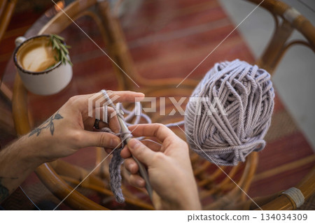 Male hands crochet handmade artwork use wool yarn and hook over table with coffee top view Male hands crochet handmade artwork use wool yarn and hook over table with coffee top view 134034309