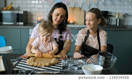 Mother guides her young daughter as she learns to handle ginger cookie dough while the older daughter helps with baking nearby Mother guides her young daughter as she learns to handle ginger cookie dough while the older daughter helps with baking nearby 134034508