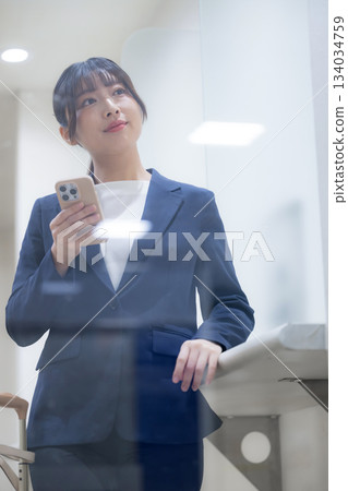 一位職業婦女站著，透過玻璃窗看著她的智慧型手機，展現了商務旅行和資訊確認的場景，垂直螢幕 134034759