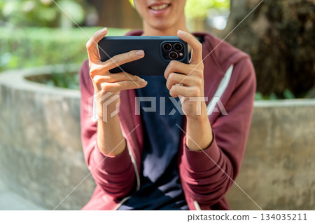 A man watching video or playing game on smartphone holding horizontally with both hands in the park. 134035211