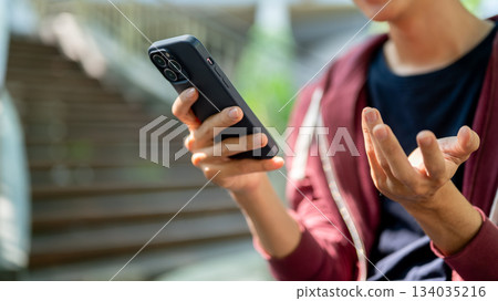 Close up of a man looking at smartphone with doing explaining gesture while sitting near the stair. 134035216