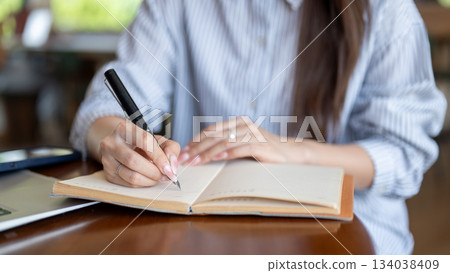 Close up of blue shirt woman writing in notebook by fountain pen while sitting at cafe wooden table Close up of blue shirt woman writing in notebook by fountain pen while sitting at cafe wooden table 134038409