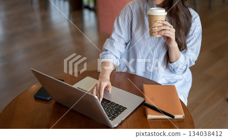 Woman in blue shirt typing on laptop while holding coffee cup and sitting at wooden table in cafe. 134038412