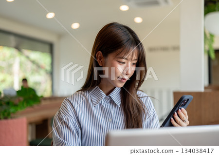 Asian woman in blue striped shirt looking at phone while sitting in front of laptop at table in cafe Asian woman in blue striped shirt looking at phone while sitting in front of laptop at table in cafe 134038417