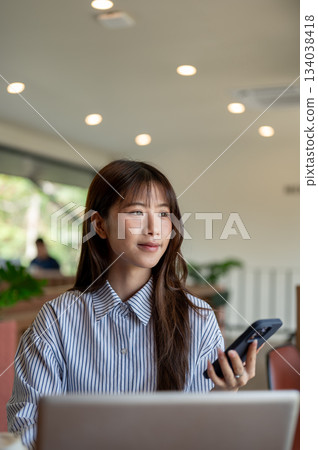 Asian woman in blue striped shirt looking away from phone while sitting in front of laptop in cafe. 134038418
