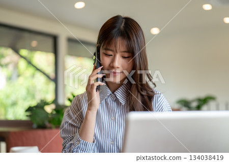 Asian woman in blue striped shirt talking on phone while sitting in front of laptop at table in cafe 134038419