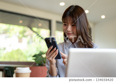 Blue striped shirt asian woman looking and typing on phone while sitting in front of laptop in cafe. 134038420