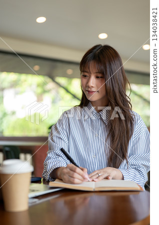Asian woman writing in notebook with fountain pen and looking at laptop while sitting in the cafe. 134038421