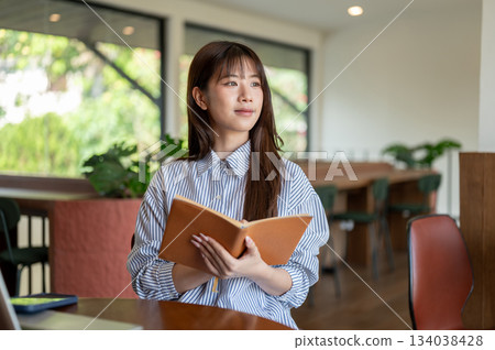 Asian woman in blue striped shirt looking away from brown book while sitting at wooden table in cafe Asian woman in blue striped shirt looking away from brown book while sitting at wooden table in cafe 134038428
